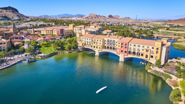 Aerial view of the Lake Las Vegas waterfront village featuring Mediterranean-style pastel buildings, lush green spaces, and serene blue water.